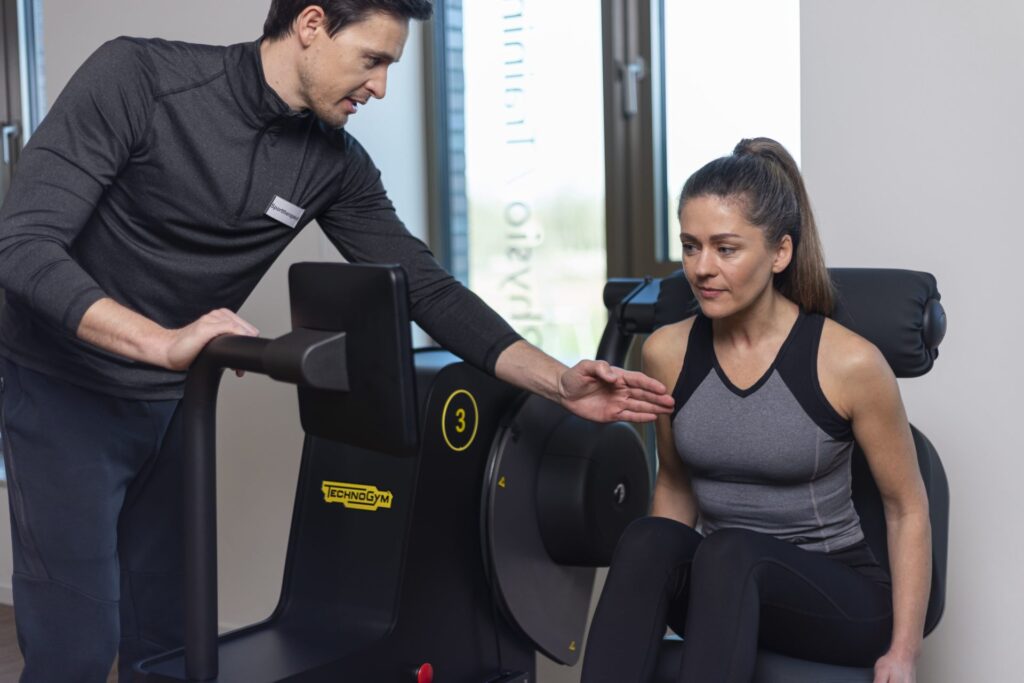 Sporttherapeut erklärt einer Frau die Bedienung eines Trainingsgeräts im Sitzen