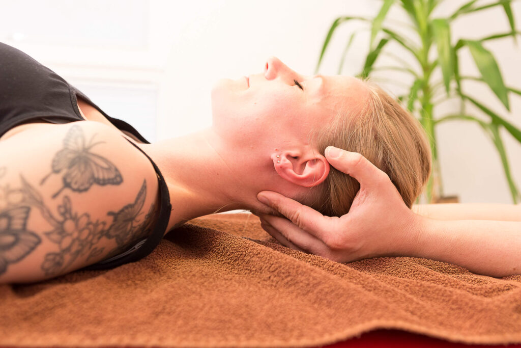 Hände stützen Kopf und Nacken einer liegenden Person auf braunem Tuch